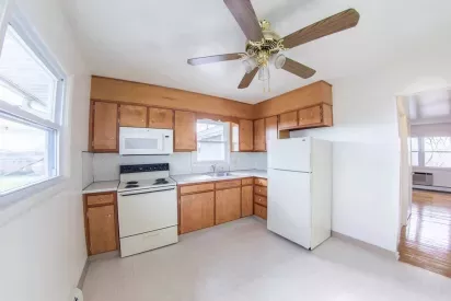 A bright kitchen with wooden cabinets, a white refrigerator, a stove, a microwave, and a ceiling fan.
