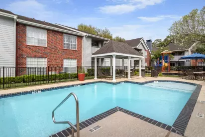 A swimming pool area with a gazebo, lounge chairs, and a children's playground surrounded by brick and siding apartment buildings.