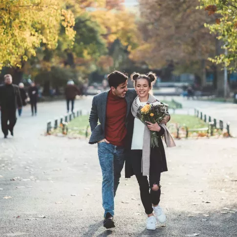 A couple walking arm-in-arm through a park in autumn, with one holding a bouquet of flowers.