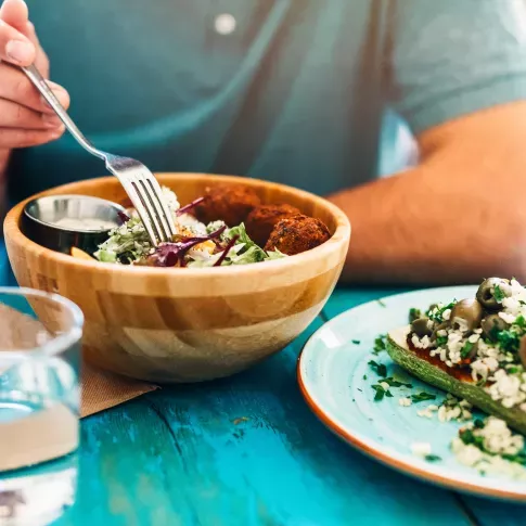 A person eating a fresh salad from a wooden bowl alongside a plate of stuffed zucchini on a vibrant blue table.
