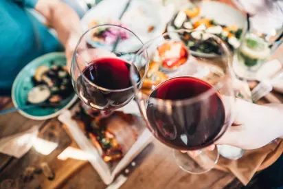 Two people toast with glasses of red wine over a table filled with colorful dishes, suggesting a lively meal shared together.