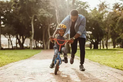 A joyful child rides a small red and blue bicycle with training wheels as a smiling adult helps guide and encourage them in a sunny park.