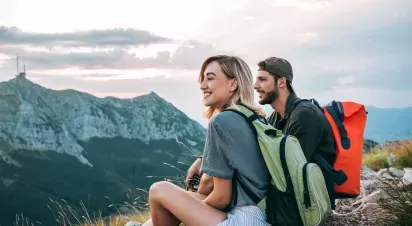 A smiling young couple with backpacks takes a break during a mountain hike, enjoying the scenic view of rugged peaks at sunset.