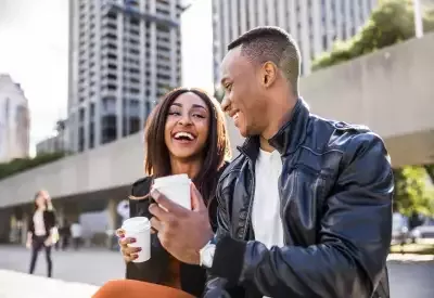 A happy couple enjoying coffee outdoors, sitting on a bench in an urban setting with tall buildings and greenery in the background, smiling and engaging in conversation.