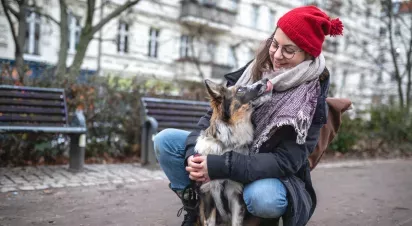 A woman wearing a red beanie kneels to embrace her dog in a park, both sharing a joyful moment.