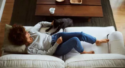 A person lounges comfortably on a couch, relaxing while their pet dog, a black French Bulldog, playfully engages with them.