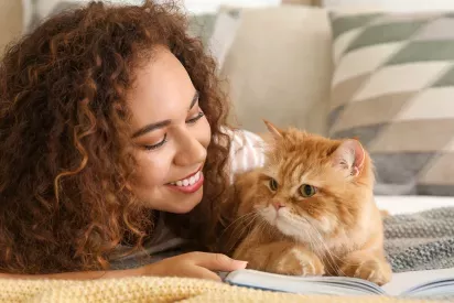 A woman with curly hair smiles warmly at a fluffy orange cat lying beside her on a cozy bed.