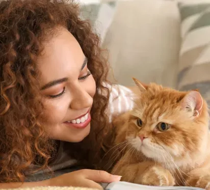 Woman smiling while lying down next to her fluffy orange cat, enjoying a peaceful moment together with a joyful and affectionate expression.