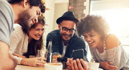 A group of friends enjoying a lighthearted moment together at a café, laughing and sharing a phone screen.