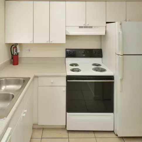 A clean and compact kitchen featuring white cabinetry, a double stainless steel sink, an electric stovetop and oven, and a refrigerator, with tiled flooring and a countertop workspace.
