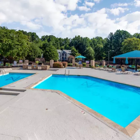 An inviting outdoor pool area with two sparkling blue pools surrounded by lounge chairs, umbrellas, and lush greenery in a serene residential setting.