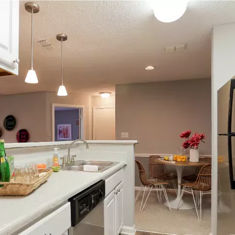 A modern kitchen setup featuring white cabinets, pendant lights, and a cozy dining area with wicker chairs and a table adorned with flowers and fruits.