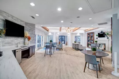 A spacious community lounge featuring modern furniture, wood-beam accents, multiple seating areas, a mounted TV, and bright natural lighting.