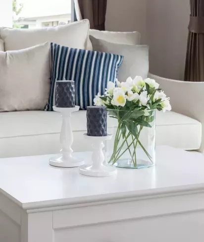A cozy living room with a white coffee table featuring two dark candles on white holders and a vase of fresh white flowers, set against a soft beige couch with striped pillows.
