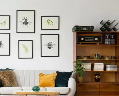 A cozy living room with a white sofa adorned with green and yellow pillows, framed botanical prints on the wall, and a wooden bookshelf displaying plants, books, and vintage decor items.