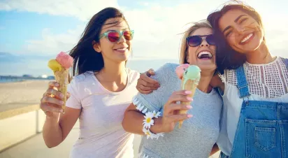 Three friends enjoying ice cream cones and laughing together at the beach.
