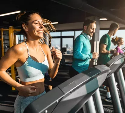 A group of individuals working out on treadmills in a brightly lit gym, featuring large windows and a variety of fitness equipment in the background.