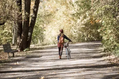 A woman walks her bicycle along a scenic, tree-lined path during autumn, surrounded by colorful foliage.