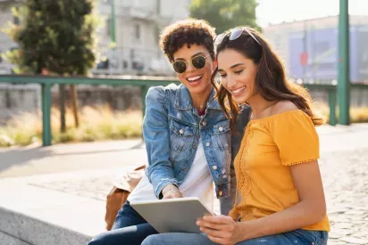 Two women are happily sitting outdoors, sharing a moment while looking at a tablet together on a sunny day.