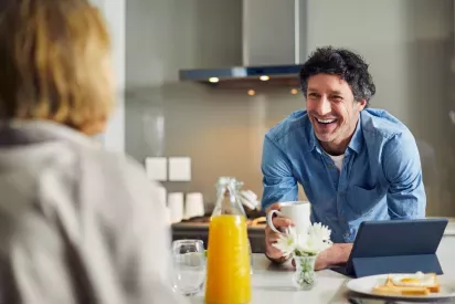 A man in a blue shirt smiles warmly while holding a coffee mug, with a breakfast setup of orange juice, flowers, and a tablet on the table.
