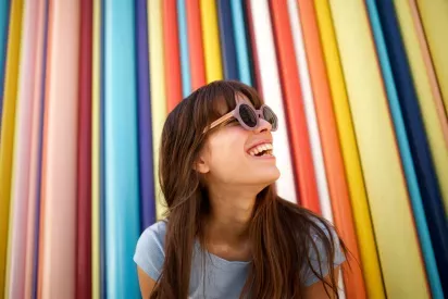 A woman wearing sunglasses smiling brightly in front of a vibrant, multicolored striped background.