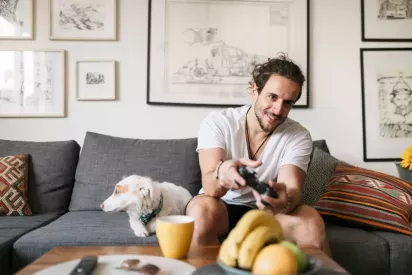 A man sits on a gray couch playing video games with his dog by his side in a cozy, art-filled living room.