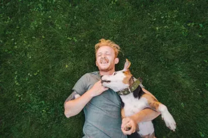A man lying on lush green grass, smiling while playing with his dog.