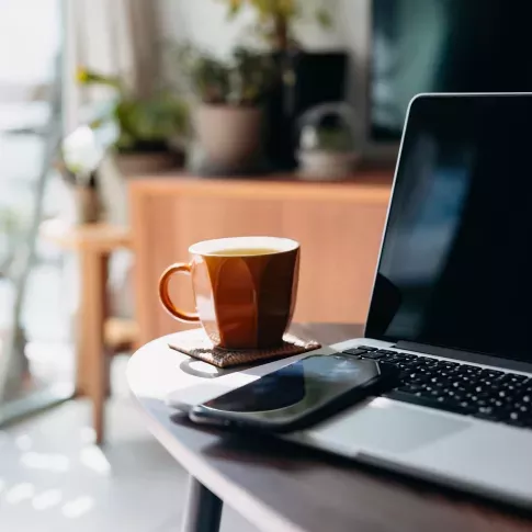 A cozy home workspace with a laptop, smartphone, and a cup of tea on a wooden table, surrounded by natural light and indoor plants in the background.