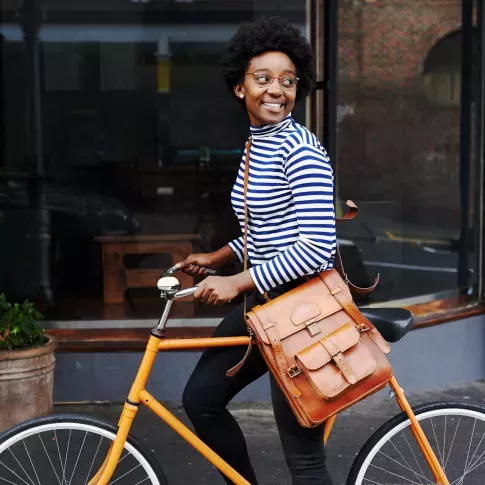 A smiling woman wearing a striped top and glasses rides a bright yellow bicycle, with a stylish leather satchel over her shoulder, in an urban setting.