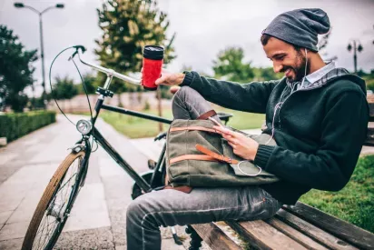 A young man sitting on a park bench with a bicycle nearby, holding a red travel mug while smiling at his phone.