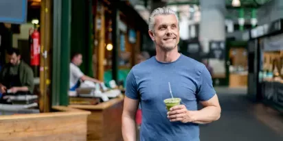 A man enjoying a green smoothie while walking through a vibrant market, smiling and relaxed.