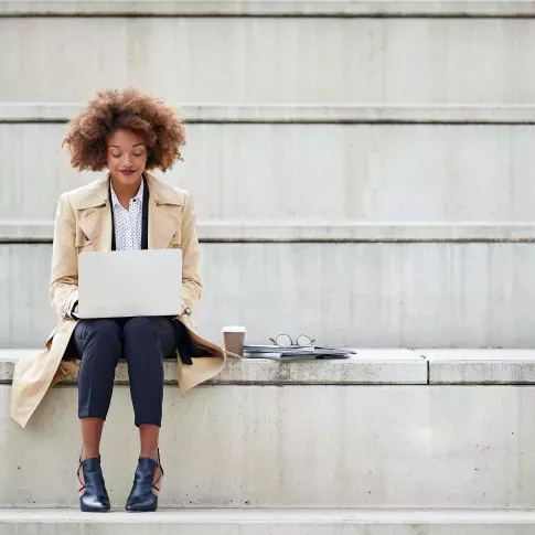 A professional woman sitting on outdoor concrete steps, working on a laptop with a coffee cup, glasses, and a notebook beside her, representing productivity and focus.