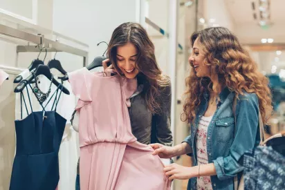 Two women joyfully shopping for clothes, with one holding a light pink dress while the other admires it.