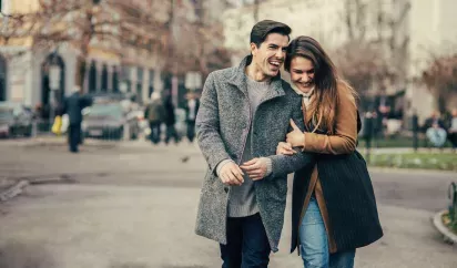 A cheerful couple walking arm in arm through a city park on a crisp day, surrounded by blurred urban scenery and trees.