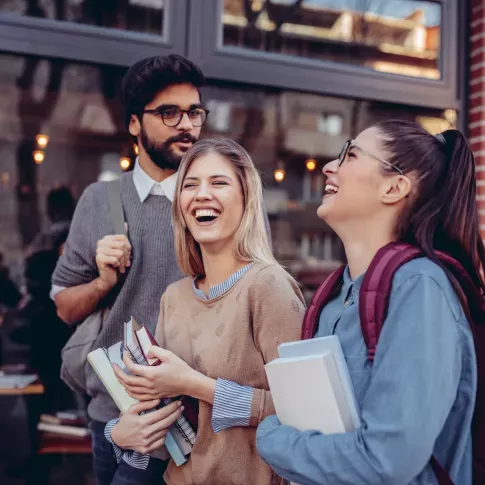 Three young adults laughing and enjoying their time together outdoors, with books and backpacks, standing near a building with large windows.