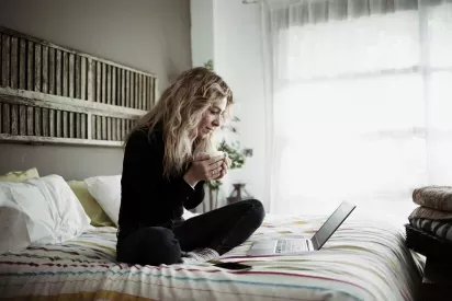 Woman sitting on bed with laptop holding coffee