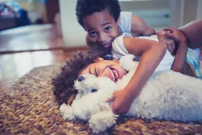 Two children happily cuddle and play with a fluffy white dog on a cozy rug in a brightly lit home.