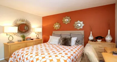  A stylish bedroom at Norcross Crossings Apartments features a bold orange accent wall, elegant decor with sunburst mirrors, and cozy furnishings for a warm and inviting atmosphere.