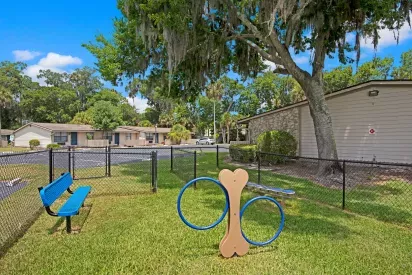 A fenced pet park with a bench and play equipment, surrounded by greenery.