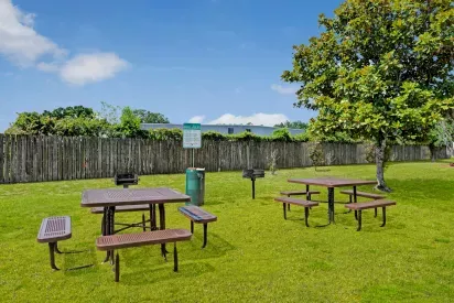 A picnic area with multiple tables and grills surrounded by a wooden fence and greenery.