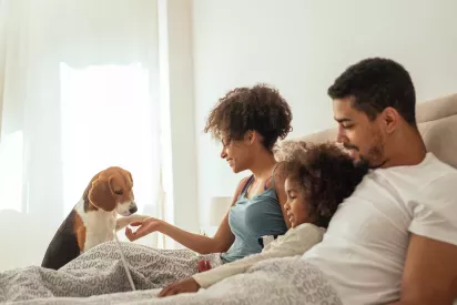 A family of three, including a young child, enjoys a relaxing moment in bed with their pet dog, smiling and engaging with each other in a cozy setting.