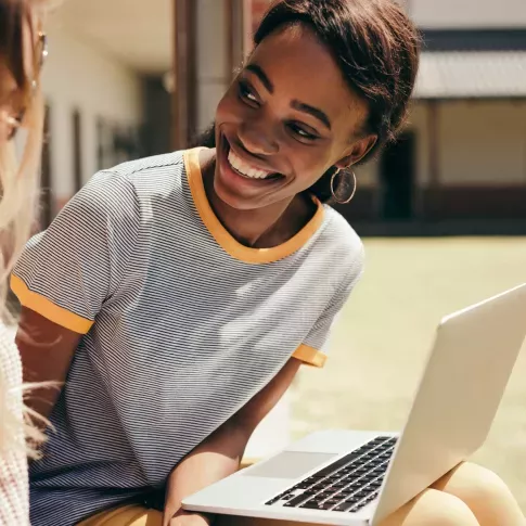 Two young women sitting outdoors, one with a laptop, smiling and engaging in conversation, highlighting the apartment community's proximity to educational opportunities and collaborative spaces.