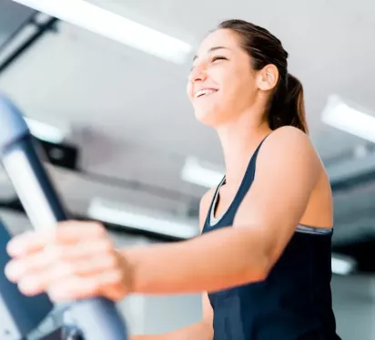 A woman smiling while using an elliptical machine in a gym, enjoying her workout in a bright, modern fitness environment.