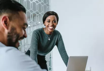 Two colleagues are engaged in a meeting, with the woman smiling as she stands near a laptop while the man is seated, smiling in the background.