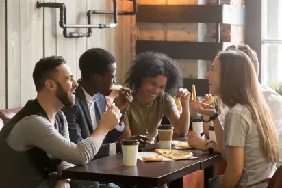 Residents of The Hamptons at Town Center in Germantown, MD, enjoying a lively conversation and pizza in a cozy, industrial-style cafe with natural light and warm decor.