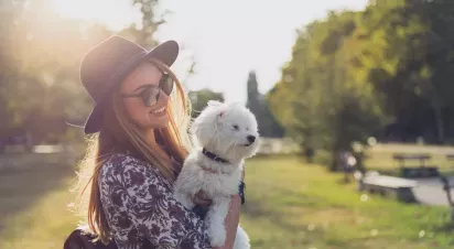A woman in sunglasses and a hat holds a small white dog in a sunny park setting with trees and benches in the background.