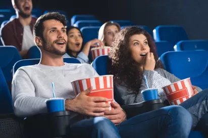 Enthusiastic moviegoers enjoying a film in a cinema, with popcorn and drinks in hand, seated in blue chairs.