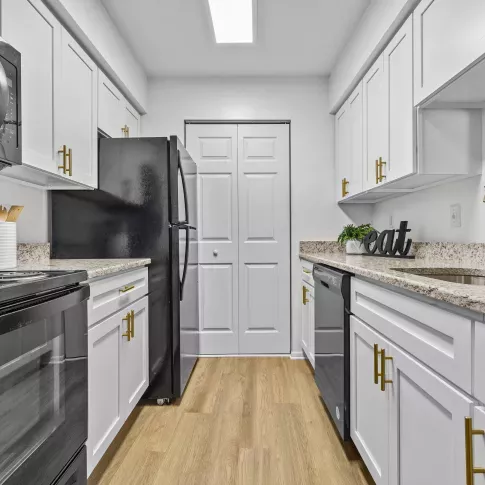 A modern galley-style kitchen featuring white cabinetry with gold handles, granite countertops, black appliances, and a stylish gold faucet, illuminated by a bright overhead light.