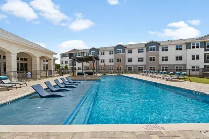 A sparkling resort-style swimming pool at Linden Ranch Luxury Apartments, featuring in-water lounge chairs and ample seating on the surrounding sun deck. The modern clubhouse and outdoor amenities provide a vibrant backdrop for relaxation.