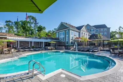 Resort-style swimming pool at Vista At Town Green Luxury Apartments, featuring crystal-clear water, shaded lounge chairs, and a clubhouse with modern architecture in the background.